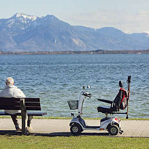 Back view of older man sitting on a bench on the waterfront with an electric wheelchair by his side.